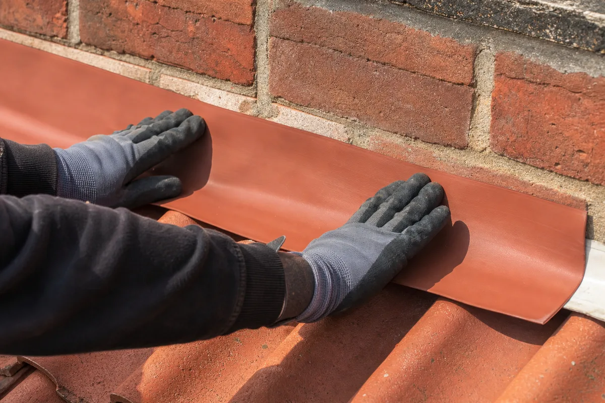 Bande souple d’étanchéité couleur terre cuite marouflée à la main contre un mur de briques, au-dessus d’un rang de tuiles
